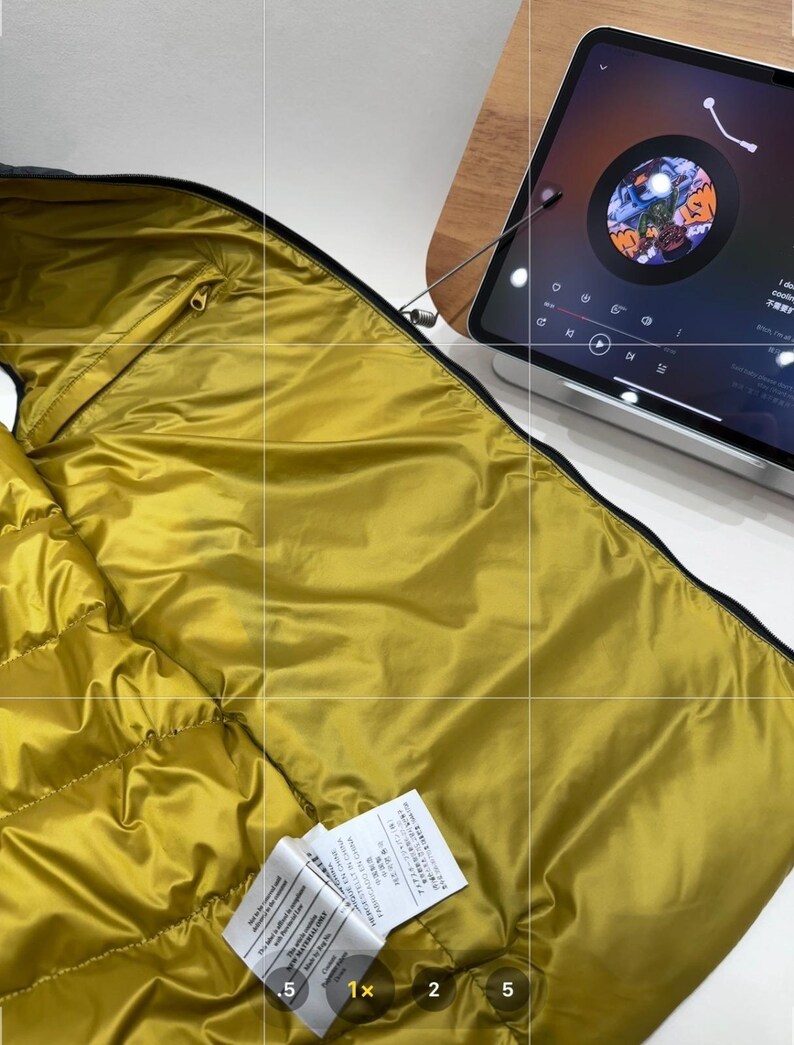 May include: Close-up of a mustard-yellow puffer jacket interior with black trim and a gold zip. The jacket is partially open, showing the quilted lining. Two white tags are visible.