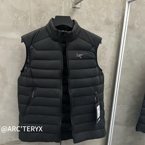May include: A black, sleeveless puffer vest with a high collar and a full-length zipper. The vest has a small logo on the chest and two zippered pockets. The brand name, @ARC'TERYX, is visible in the lower left corner.