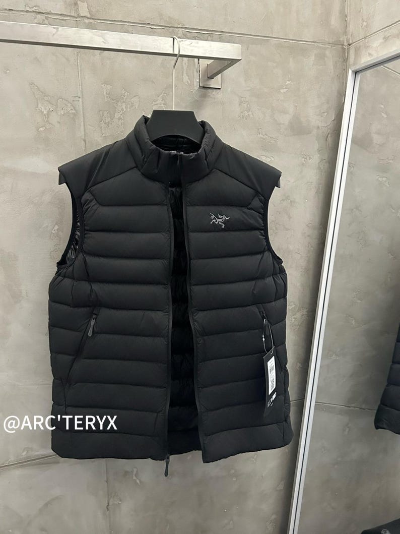 May include: A black, sleeveless puffer vest with a high collar and a full-length zipper. The vest has a small logo on the chest and two zippered pockets. The brand name, @ARC'TERYX, is visible in the lower left corner.