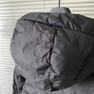 Op de afbeelding: Zwarte jas met capuchon met de merknaam "ARCTERYX" geborduurd op de capuchon. De jas is gemaakt van een gladde, matte stof en lijkt licht van gewicht. De capuchon is ontworpen om te beschermen tegen de elementen.