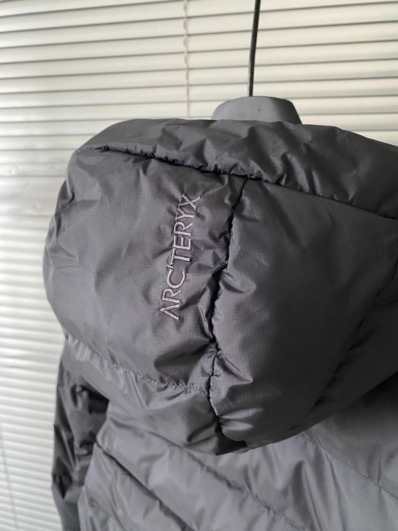 Op de afbeelding: Zwarte jas met capuchon met de merknaam "ARCTERYX" geborduurd op de capuchon. De jas is gemaakt van een gladde, matte stof en lijkt licht van gewicht. De capuchon is ontworpen om te beschermen tegen de elementen.