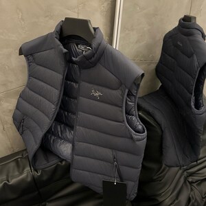 May include: A dark gray puffer vest with a high collar and a zipper closure. The vest has a quilted design and a small logo on the chest. It is hanging on a black hanger.
