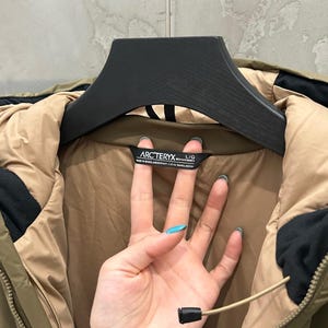 May include: Olive green and tan jacket with a black hood and a black zipper. The jacket has a black label with the brand name "ARCTERYX" printed on it. The jacket is hanging on a black hanger.