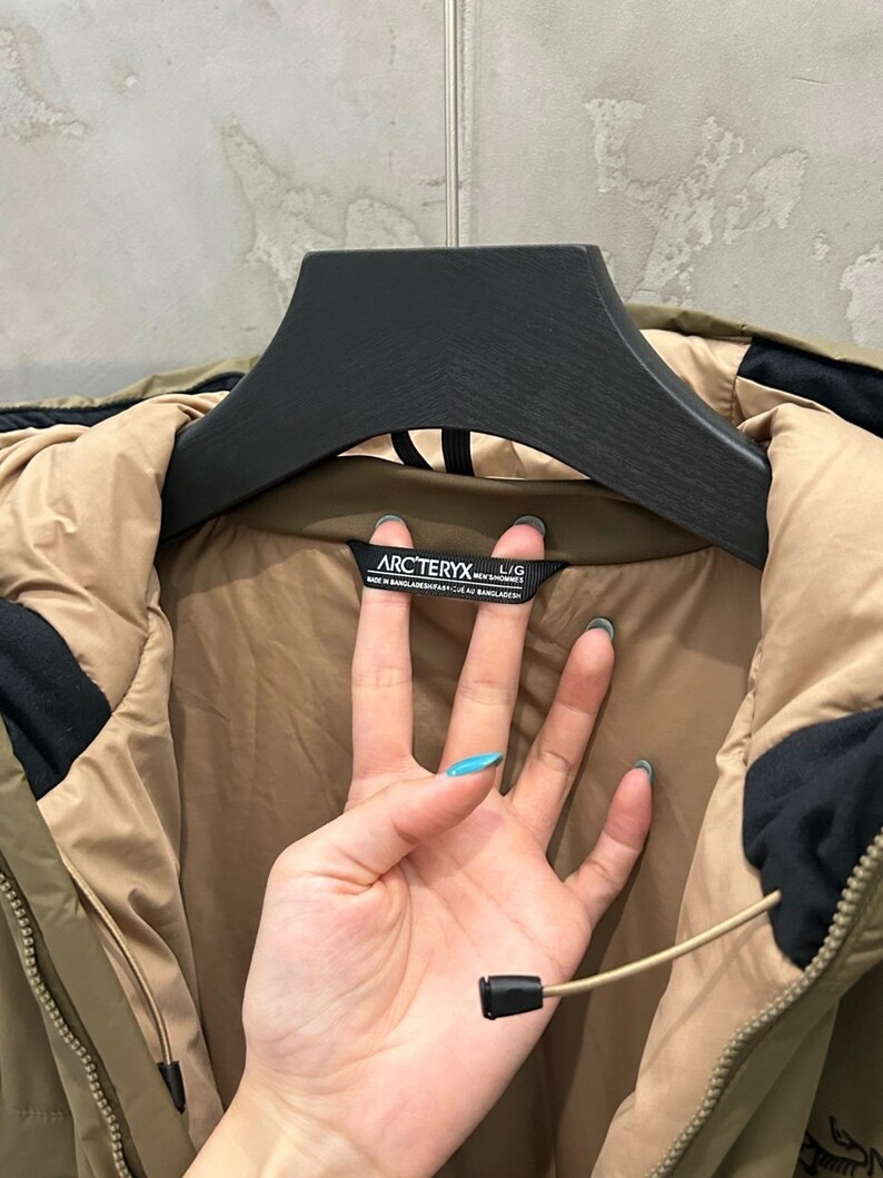 May include: Olive green and tan jacket with a black hood and a black zipper. The jacket has a black label with the brand name "ARCTERYX" printed on it. The jacket is hanging on a black hanger.