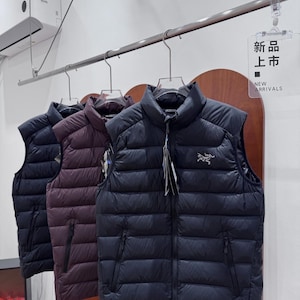 May include: Three padded gilets in navy, burgundy, and black hang on a rail. The gilets have a high collar, a zip closure, and two side pockets. The sign reads "NEW ARRIVALS".