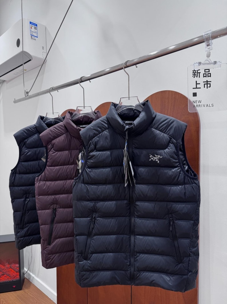 May include: Three padded gilets in navy, burgundy, and black hang on a rail. The gilets have a high collar, a zip closure, and two side pockets. The sign reads "NEW ARRIVALS".