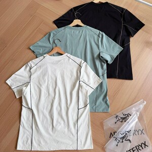 May include: Three short-sleeved t-shirts in white, teal, and black, each with black stitching details. The shirts hang on wooden hangers against a wooden floor. A clear plastic bag with the text "ARC'TERYX" is also visible.