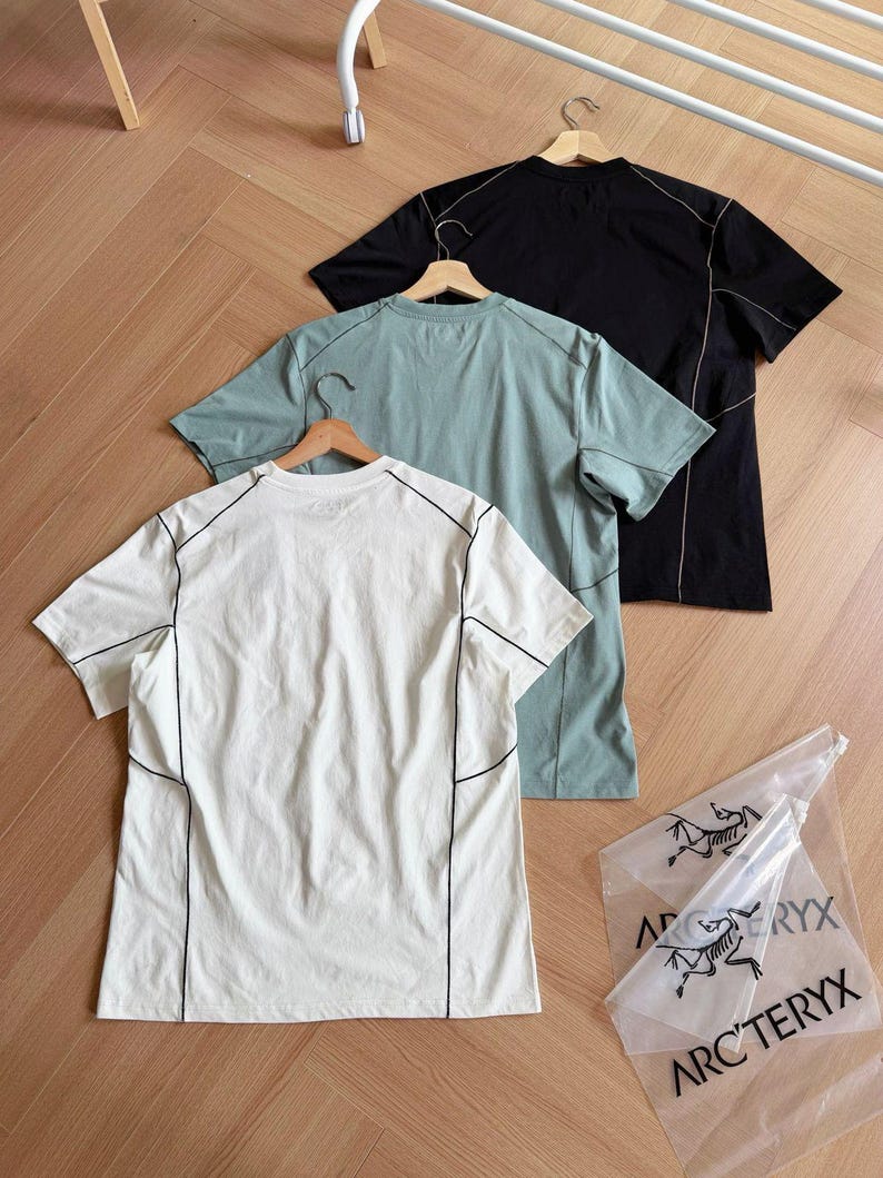 May include: Three short-sleeved t-shirts in white, teal, and black, each with black stitching details. The shirts hang on wooden hangers against a wooden floor. A clear plastic bag with the text "ARC'TERYX" is also visible.
