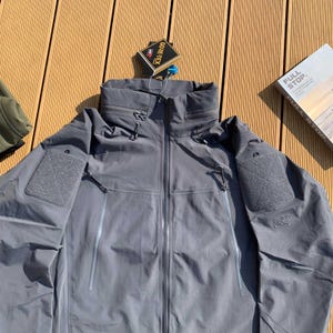 Op de afbeelding: Grijze, waterdichte jas met een hoge kraag en meerdere zakken. De jas heeft een ritssluiting en een GORE-TEX-label. De jas is ontworpen voor buitenactiviteiten en is gemaakt van duurzaam materiaal.