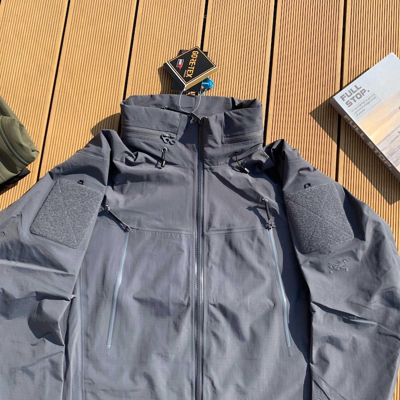 Op de afbeelding: Grijze, waterdichte jas met een hoge kraag en meerdere zakken. De jas heeft een ritssluiting en een GORE-TEX-label. De jas is ontworpen voor buitenactiviteiten en is gemaakt van duurzaam materiaal.
