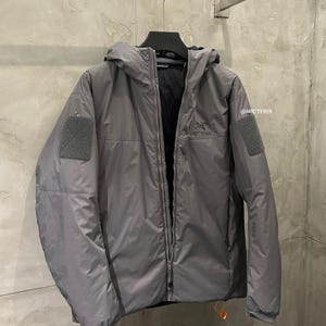 May include: A gray hooded jacket with a full-length zipper, two patch pockets, and the brand name "ARC'TERYX" printed on the chest. The jacket is hanging on a black hanger against a neutral background.