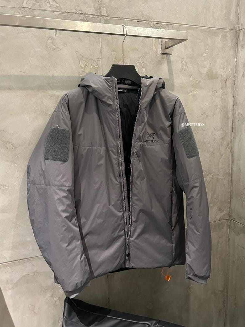 May include: A gray hooded jacket with a full-length zipper, two patch pockets, and the brand name "ARC'TERYX" printed on the chest. The jacket is hanging on a black hanger against a neutral background.
