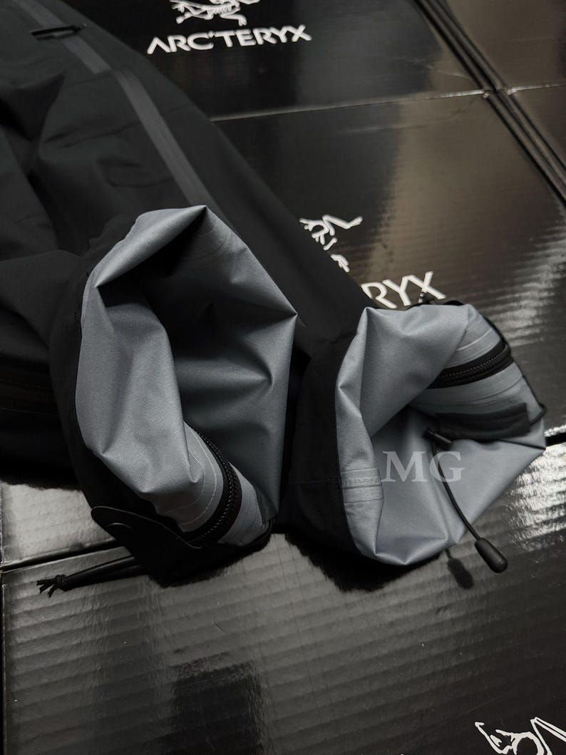 May include: Close-up of a black jacket sleeve with a gray interior lining. The sleeve has a zipper and a drawstring at the cuff. The Arc'teryx logo is visible in the background.