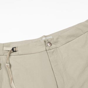 Pu&ograve; includere: Primo piano di pantaloni da esterno color beige. Presentano chiusura con bottone, passanti per cintura e tasche con cerniera. Il marchio "Arc'teryx" &egrave; visibile in vita. I pantaloni hanno un design minimalista.