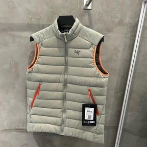 May include: A light gray puffer vest with a high collar and a full-length zipper. The vest has orange trim around the armholes and two orange zippered pockets. A black tag with white text and a QR code hangs from the zipper.