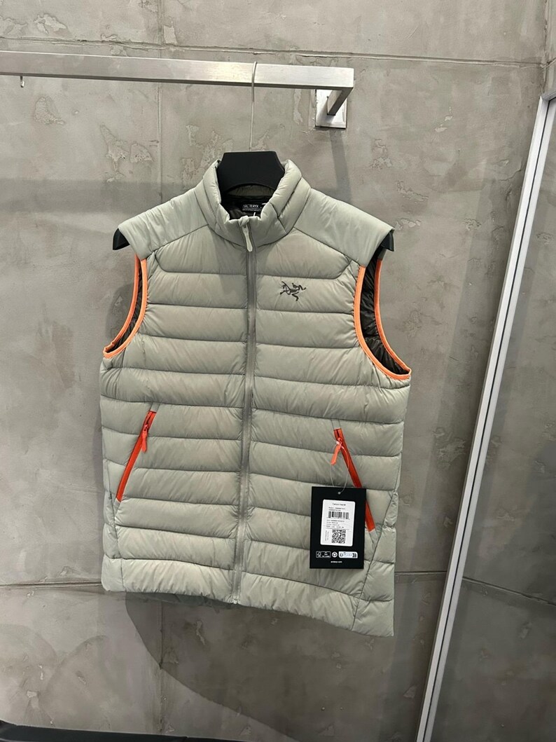 May include: A light gray puffer vest with a high collar and a full-length zipper. The vest has orange trim around the armholes and two orange zippered pockets. A black tag with white text and a QR code hangs from the zipper.