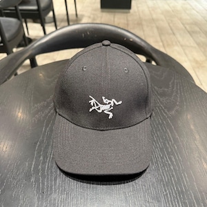 Puede incluir: Gorra de b&eacute;isbol negra con visera curva. La gorra presenta un logotipo blanco en la parte delantera y un bot&oacute;n en la parte superior. Descansa sobre una superficie de madera oscura.