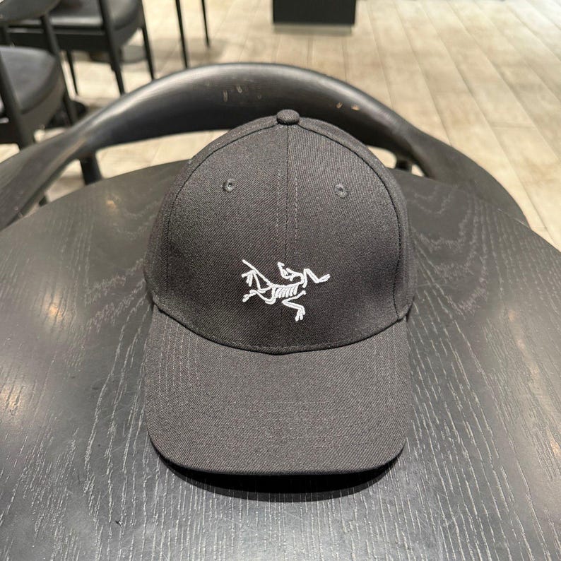 Puede incluir: Gorra de b&eacute;isbol negra con visera curva. La gorra presenta un logotipo blanco en la parte delantera y un bot&oacute;n en la parte superior. Descansa sobre una superficie de madera oscura.