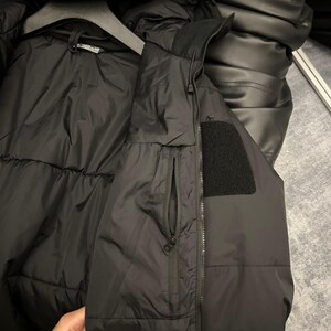 May include: Black puffer jacket with a high collar and a zippered pocket. The jacket has a black patch on the upper arm and is hanging on a black hanger. The jacket appears to be made of a water-resistant material.
