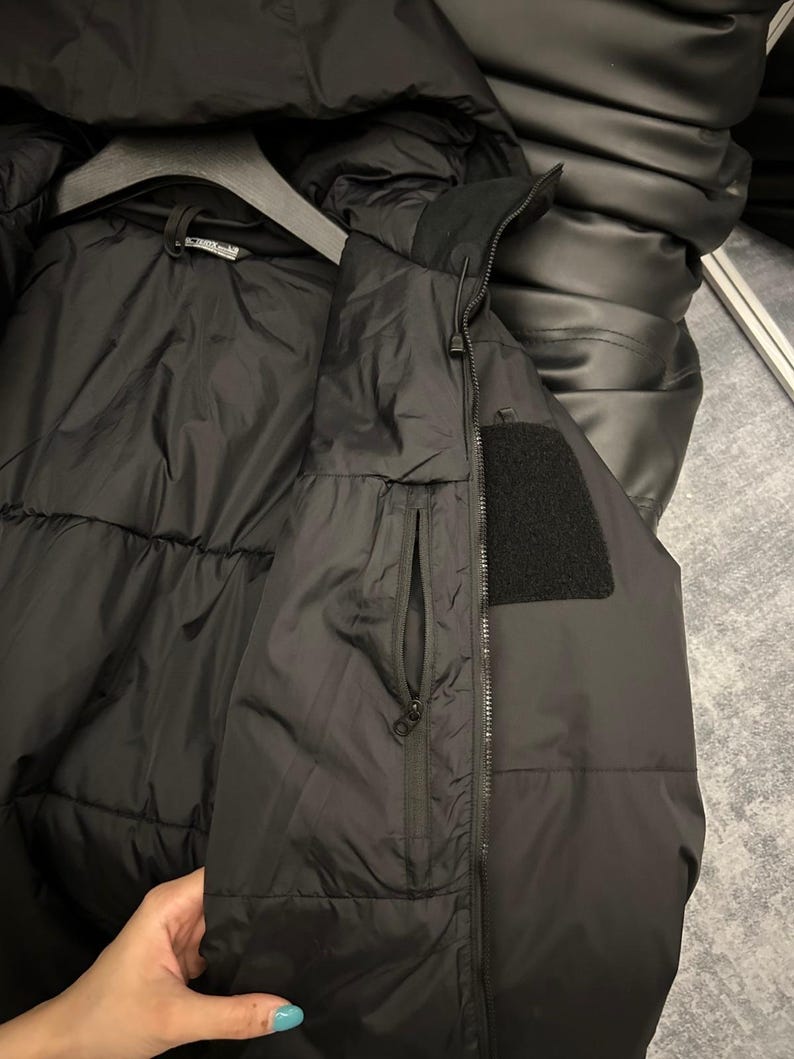 May include: Black puffer jacket with a high collar and a zippered pocket. The jacket has a black patch on the upper arm and is hanging on a black hanger. The jacket appears to be made of a water-resistant material.