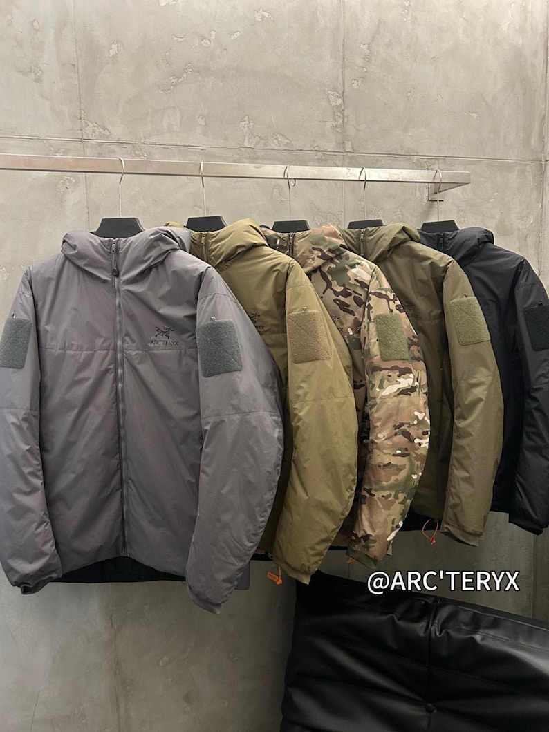 May include: A row of hooded jackets in various colors, including gray, olive green, camouflage, and black, hang on a metal rack. Each jacket has a patch on the upper arm and a zipper closure. The brand name is visible on the bottom right.