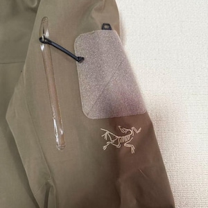 Op de afbeelding: Close-up van een kaki jas mouw. Voorzien van een zak met ritssluiting, een klittenband patch en een kleine zwarte lus. Het Arc'teryx logo is wit geborduurd op de onderste mouw.