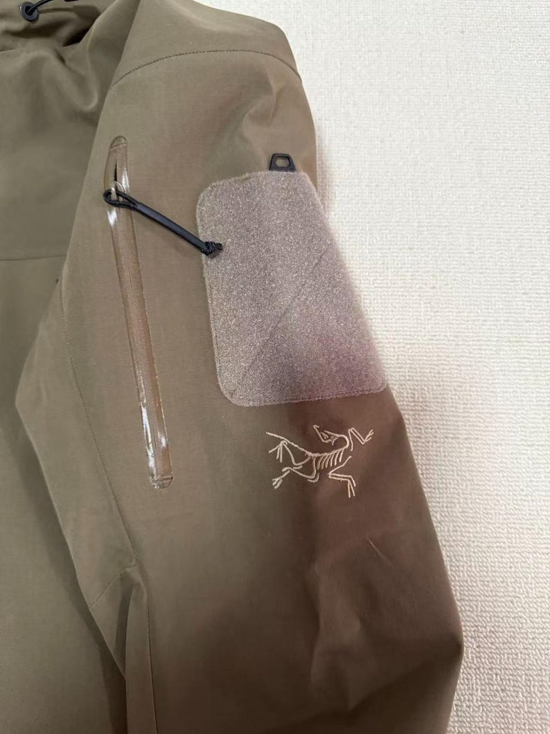 Op de afbeelding: Close-up van een kaki jas mouw. Voorzien van een zak met ritssluiting, een klittenband patch en een kleine zwarte lus. Het Arc'teryx logo is wit geborduurd op de onderste mouw.