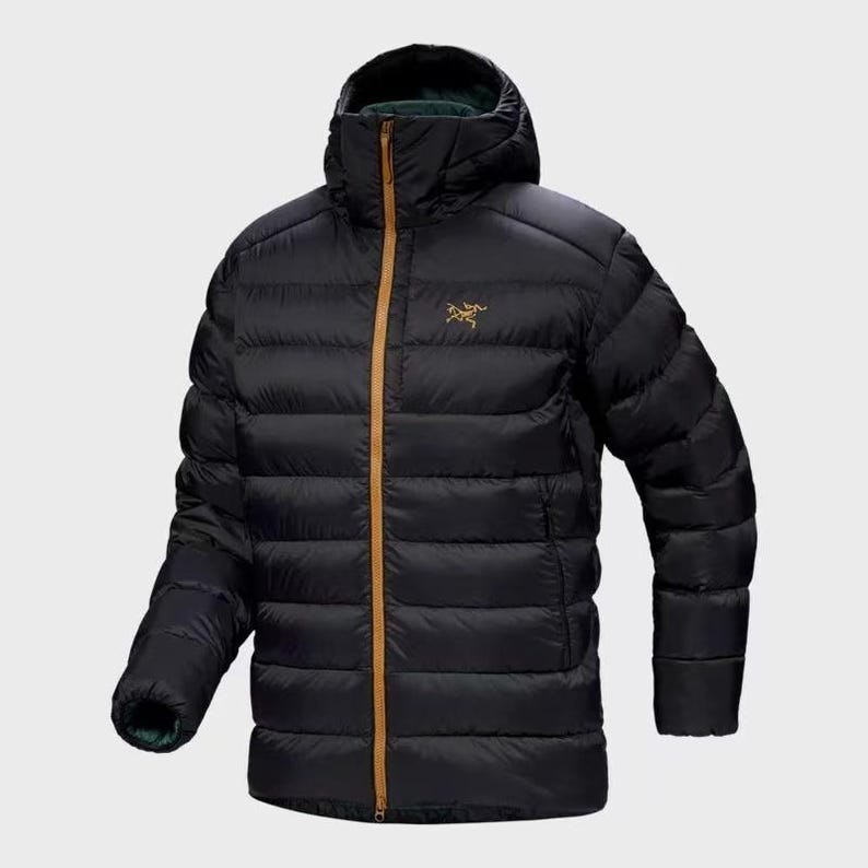Op de afbeelding: Zwarte gewatteerde jas met capuchon, gouden rits en logo. De jas heeft een gewatteerd ontwerp en een hoge kraag. Ontworpen voor koud weer en buitenactiviteiten.