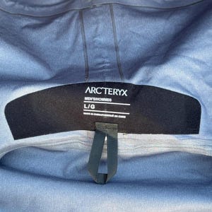 May include: Close-up of a gray jacket interior with a black label. The label reads "ARC'TERYX" in white text, with "MEN'S/HOMMES" and "L/G" below. The jacket is made in China.