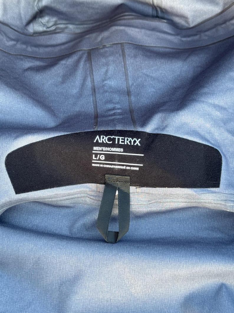 May include: Close-up of a gray jacket interior with a black label. The label reads "ARC'TERYX" in white text, with "MEN'S/HOMMES" and "L/G" below. The jacket is made in China.