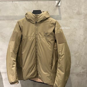 May include: A tan-colored hooded jacket with a full-length zipper and the brand name "Arc'teryx" on the chest. The jacket has two patch pockets on the sleeves and an orange pull tab on the zipper. The jacket is hanging on a metal bar.