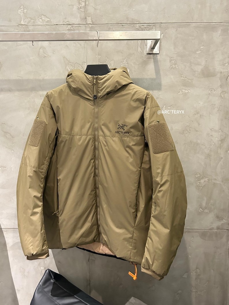 May include: A tan-colored hooded jacket with a full-length zipper and the brand name "Arc'teryx" on the chest. The jacket has two patch pockets on the sleeves and an orange pull tab on the zipper. The jacket is hanging on a metal bar.