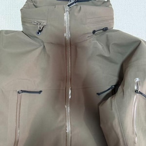 Op de afbeelding: Een bruinbeige jas met een lange ritssluiting en een hoge kraag. De jas heeft meerdere zakken en trekkoorden. Een label met de merknaam "Arc'teryx" is bevestigd.