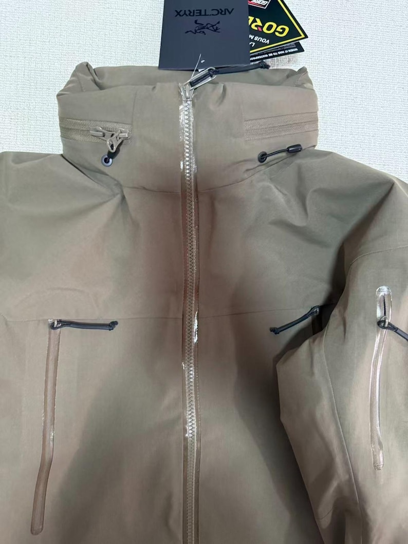 Op de afbeelding: Een bruinbeige jas met een lange ritssluiting en een hoge kraag. De jas heeft meerdere zakken en trekkoorden. Een label met de merknaam "Arc'teryx" is bevestigd.