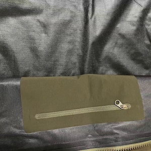 Op de afbeelding: Close-up van de binnenkant van een donkergrijze jas. Een rechthoekige olijfgroene zak met een rits is op de voering van de jas genaaid. De rits is goudkleurig en de voering van de jas heeft een getextureerde uitstraling.