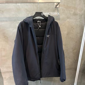 Op de afbeelding: Een marineblauwe jas met capuchon en ritssluiting, hangend aan een zwarte hanger. De jas heeft een gewatteerde binnenkant en een klein logo op de borst. De tekst "@ARC'TERYX" is zichtbaar.