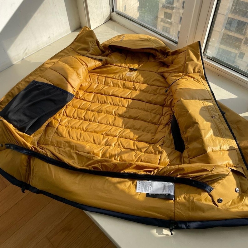 May include: Mustard-yellow puffer jacket with a hood, open to show the quilted interior. The jacket features a black pocket and trim. It is lying on a wooden surface near a window.