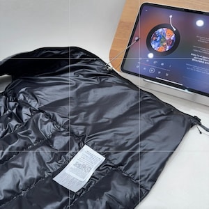 May include: A black, quilted puffer jacket with a glossy finish. The jacket is partially unzipped, revealing a white label with printed text. A tablet displaying a colourful image is in the background.