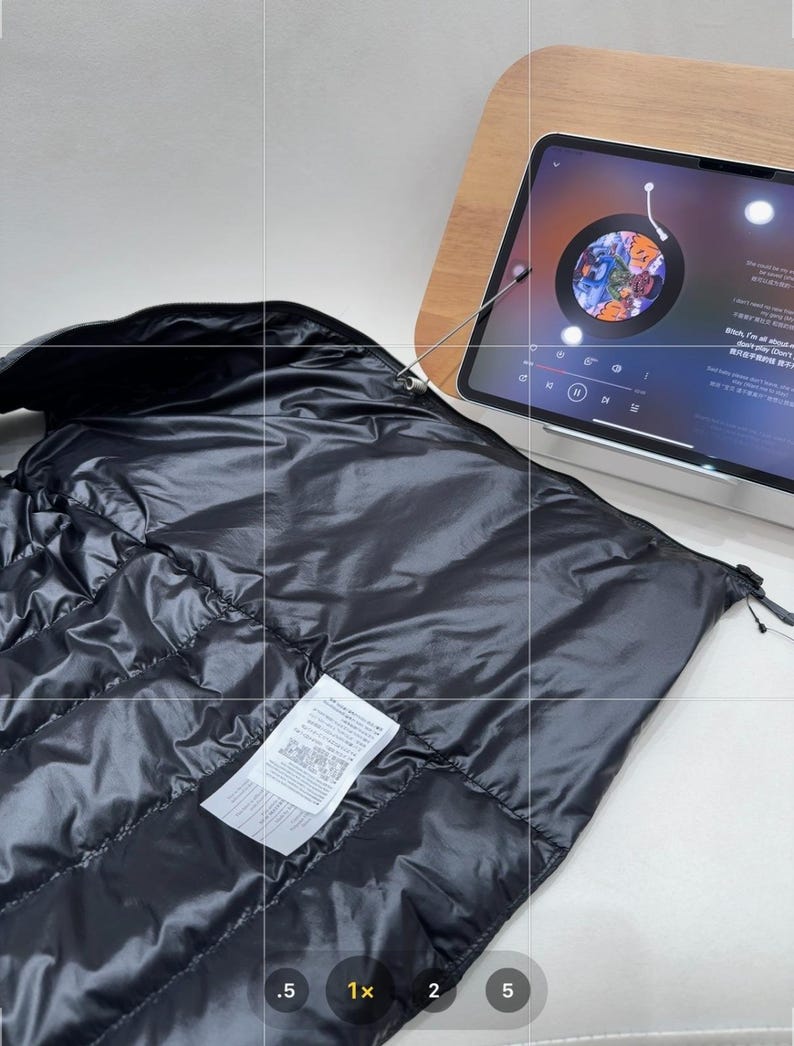 May include: A black, quilted puffer jacket with a glossy finish. The jacket is partially unzipped, revealing a white label with printed text. A tablet displaying a colourful image is in the background.