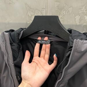 May include: A gray and black hooded jacket with a zipper closure. The jacket has a black interior and a gray exterior. The brand name "Arc'teryx" is visible on a tag inside the collar. The jacket is hanging on a black hanger.