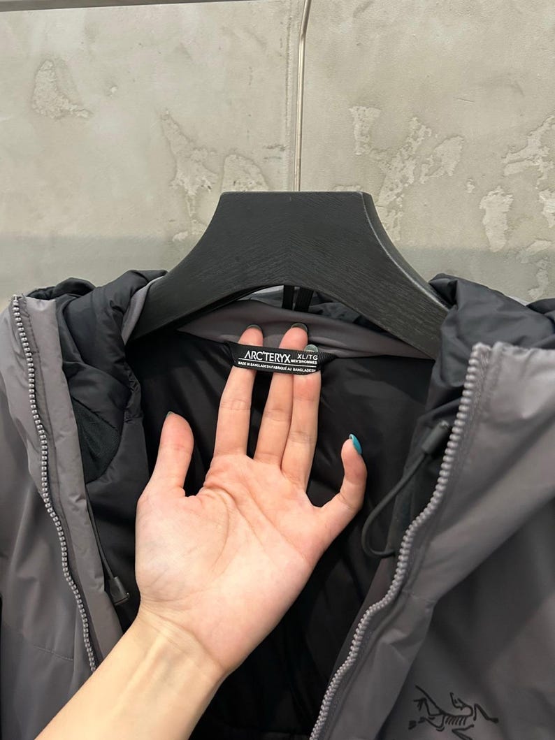 May include: A gray and black hooded jacket with a zipper closure. The jacket has a black interior and a gray exterior. The brand name "Arc'teryx" is visible on a tag inside the collar. The jacket is hanging on a black hanger.
