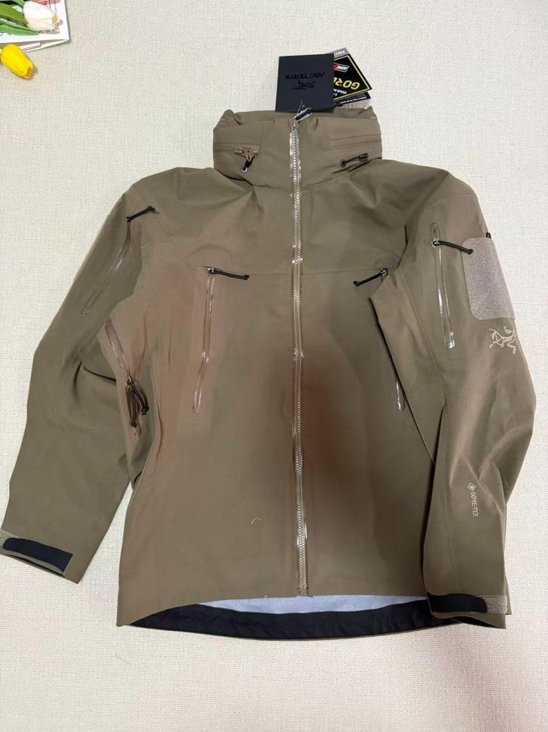 Op de afbeelding: Een bruin gekleurde outdoorjas met een hoge kraag en meerdere zakken met ritssluiting. De jas heeft een lange ritssluiting en een klein logo op de mouw. De jas is gemaakt van waterdicht materiaal en is ontworpen voor buitenactiviteiten.