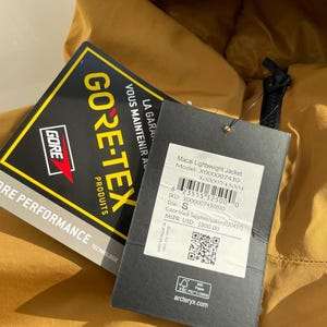 May include: Mustard-coloured Macai Lightweight Jacket with a black tag. The tag displays the GORE-TEX logo and product information, including the model number, size S, and colour black Sapphire/yukon.