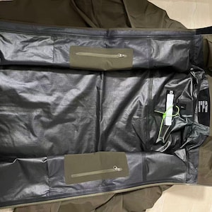 Op de afbeelding: Een olijfgroene jas met een zwarte binnenkant. De jas heeft twee olijfgroene zakken met ritsen en een zwart label met witte tekst. De binnenkant is van glanzend, waterdicht materiaal. Een wit label met groen touw is bevestigd.