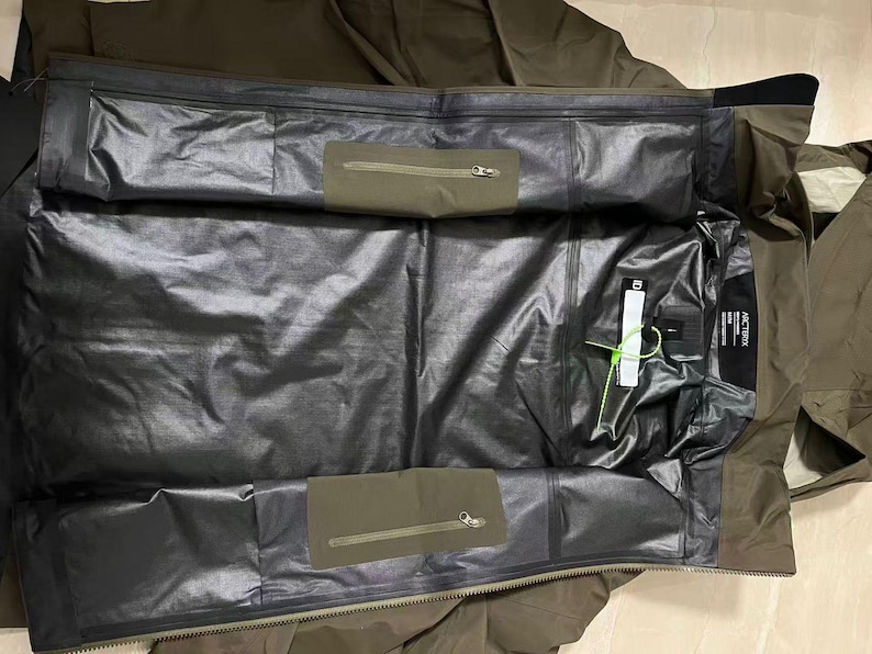 Op de afbeelding: Een olijfgroene jas met een zwarte binnenkant. De jas heeft twee olijfgroene zakken met ritsen en een zwart label met witte tekst. De binnenkant is van glanzend, waterdicht materiaal. Een wit label met groen touw is bevestigd.