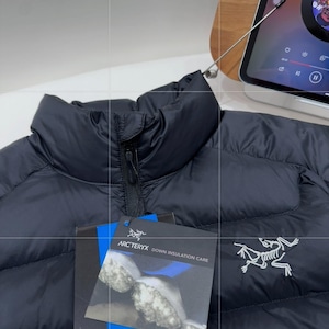 May include: A black down jacket with a high collar and a zip fastening. The jacket features the Arc'teryx logo and a tag with "Arc'teryx Down Insulation Care". The jacket is made of a smooth, quilted material.