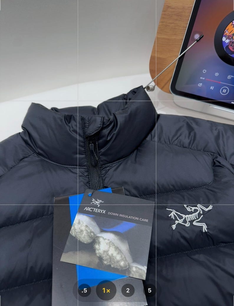 May include: A black down jacket with a high collar and a zip fastening. The jacket features the Arc'teryx logo and a tag with "Arc'teryx Down Insulation Care". The jacket is made of a smooth, quilted material.