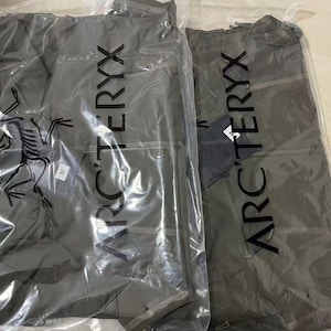 Op de afbeelding: Twee grijze jassen in doorzichtige plastic verpakking. De jassen hebben de merknaam "ARC'TERYX" verticaal op de verpakking gedrukt. E&eacute;n jas heeft een label. De jassen zijn opgevouwen en zien er nieuw uit.