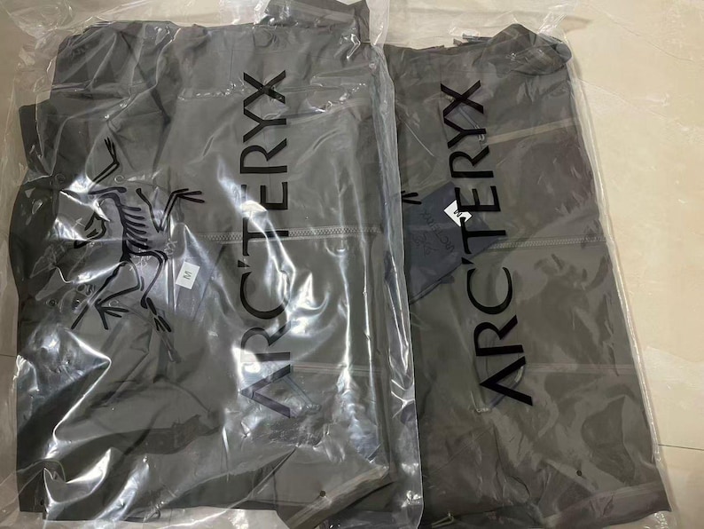 Op de afbeelding: Twee grijze jassen in doorzichtige plastic verpakking. De jassen hebben de merknaam "ARC'TERYX" verticaal op de verpakking gedrukt. E&eacute;n jas heeft een label. De jassen zijn opgevouwen en zien er nieuw uit.