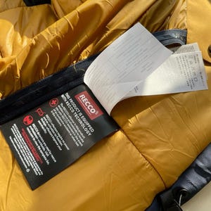 May include: Close-up of a mustard yellow jacket interior with a black label that reads "THIS PRODUCT IS EQUIPPED WITH RECCO TECHNOLOGY". The label describes the RECCO reflector technology used in the jacket for rescue purposes.
